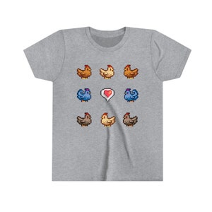 Pu&ograve; includere: Una maglietta grigia con un design a pixel di galline. Il design presenta un cuore al centro e diverse galline di diversi colori, tra cui marrone, giallo e blu.