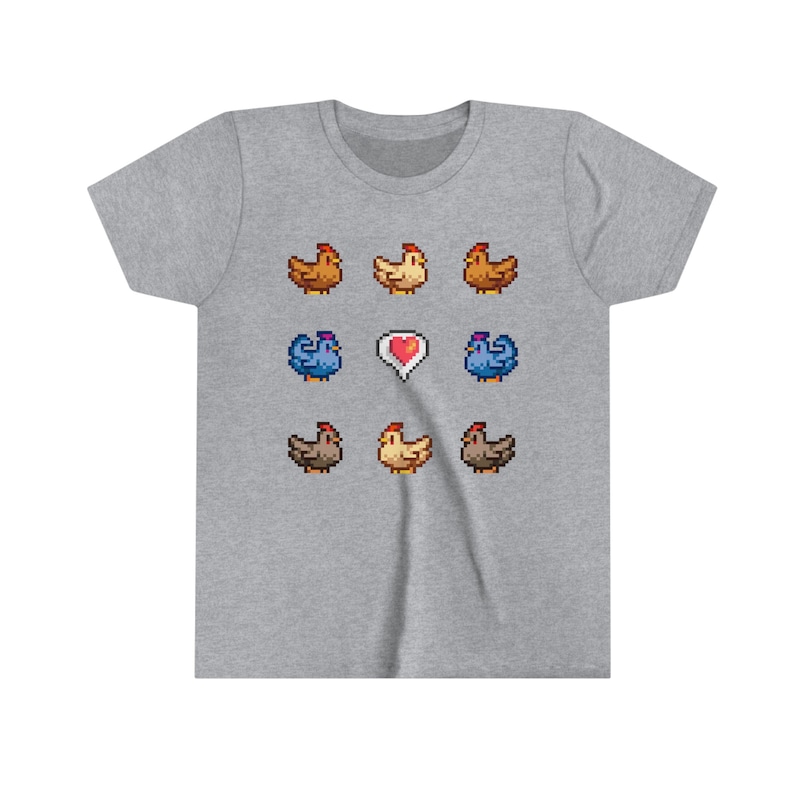 Pu&ograve; includere: Una maglietta grigia con un design a pixel di galline. Il design presenta un cuore al centro e diverse galline di diversi colori, tra cui marrone, giallo e blu.