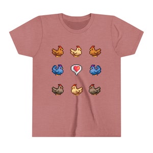 Pu&ograve; includere: Una maglietta rosa con un design a pixel di nove galline e un cuore. Le galline sono marroni, gialle e blu.