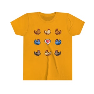 Pu&ograve; includere: Una maglietta gialla con un design a pixel di galline e un cuore. Le galline sono marroni, bianche e blu.