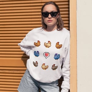 May include: White sweatshirt featuring a pixelated design of chickens and a heart. The chickens are in various colours, including brown, yellow, and blue.