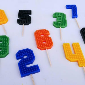 Op de afbeelding: Assortiment van kleurrijke nummerkaarsen met een getextureerd baksteenpatroon. De kaarsen zijn in de kleuren zwart, groen, oranje, geel, blauw en rood. Elke kaars zit op een houten stokje.