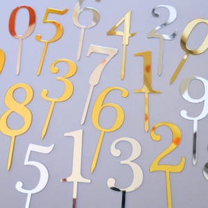 Op de afbeelding: Een verzameling reflecterende taarttoppers in de vorm van de cijfers nul tot en met negen, met metallic gouden en zilveren afwerkingen. Elk cijfer is bevestigd aan een dunne, rechte prikker die bedoeld is om in voedsel te worden gestoken.