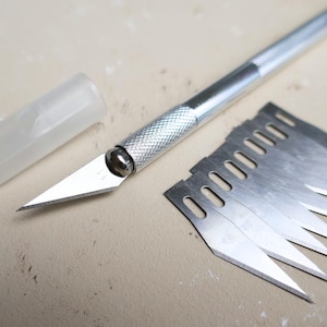 Könnte beinhalten: Ein silbernes Bastelmesser mit strukturiertem Griff und scharfer Klinge, zusammen mit einem Satz Ersatzklingen. Das Messer hat eine transparente Kunststoffklingenabdeckung. Die Klingen sind aus Stahl.