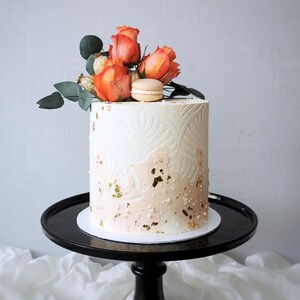 May include: A round, white cake with a textured design and peach-colored accents. The cake is decorated with orange roses, green leaves, a macaron, and gold leaf accents. It sits on a black cake stand.