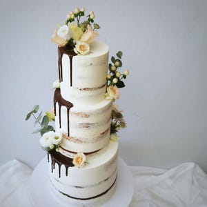 May include: A three-tiered cake with white frosting and chocolate ganache drips. The cake is decorated with fresh flowers, including roses and greenery. The cake sits on a white cake stand, perfect for a wedding or celebration.