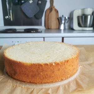 May include: A freshly baked, round cake with a golden-brown crust sits on parchment paper. The cake is light in color and appears to be a sponge cake, ready for frosting and decoration. The background shows a kitchen setting with cooking utensils.