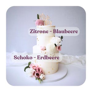May include: A three-tiered wedding cake with white frosting and gold leaf accents. The cake is decorated with pink and white flowers and a wooden "Mr & Mrs" cake topper. The text "Zitrone - Blaubeere" and "Schoko - Erdbeere" are visible.
