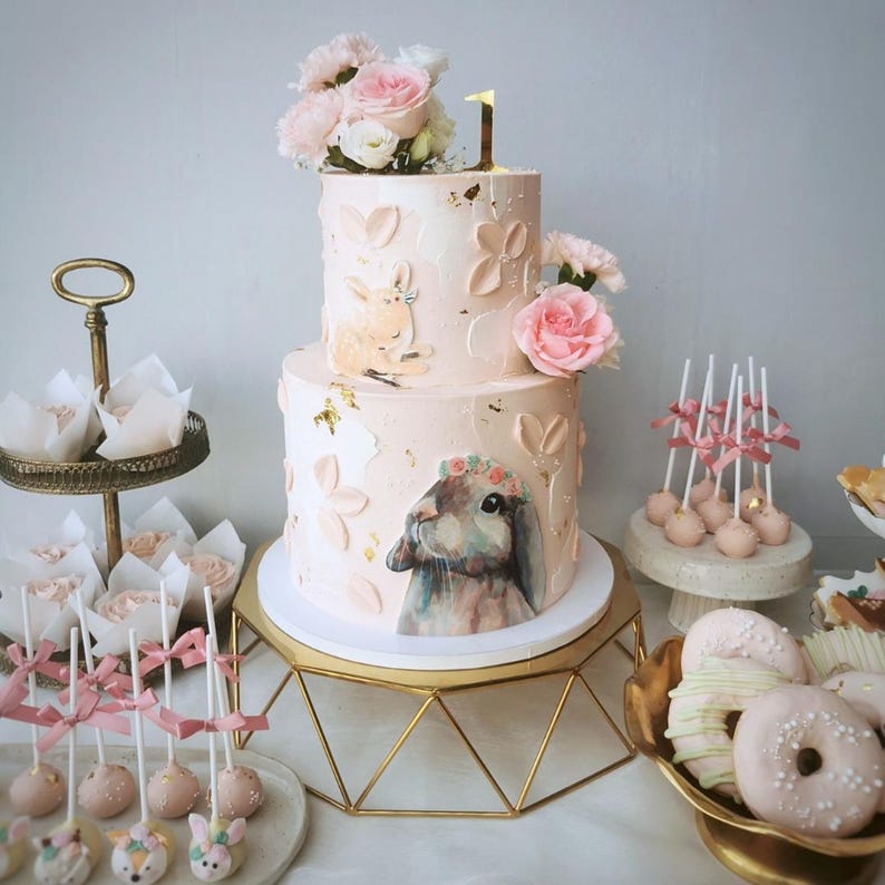 May include: A two-tiered pink cake decorated with floral accents, gold leaf, and a bunny illustration. The cake is topped with fresh flowers and a gold number 1. Other treats include cake pops, cupcakes, and donuts.