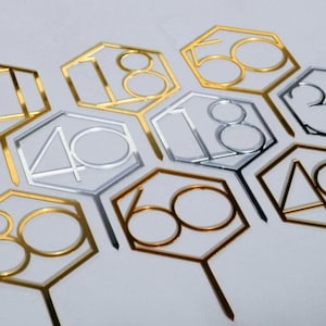 Op de afbeelding: Een set gouden en zilveren zeshoekige taarttoppers met de nummers 18, 40, 60, 18, 50, 30 en 40.