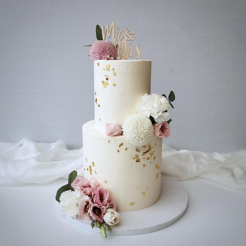 May include: A two-tiered white wedding cake decorated with gold leaf accents and fresh flowers. The cake is topped with a wooden "Mr. & Mrs." cake topper. Pink and white flowers adorn the cake's base and top tier.
