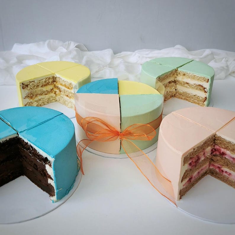 May include: Five small, round cakes with colorful frosting and cut into quarters. The cakes have layers of filling and are decorated with blue, yellow, green, and pink frosting. One cake is tied with an orange ribbon. The cakes sit on white surfaces.