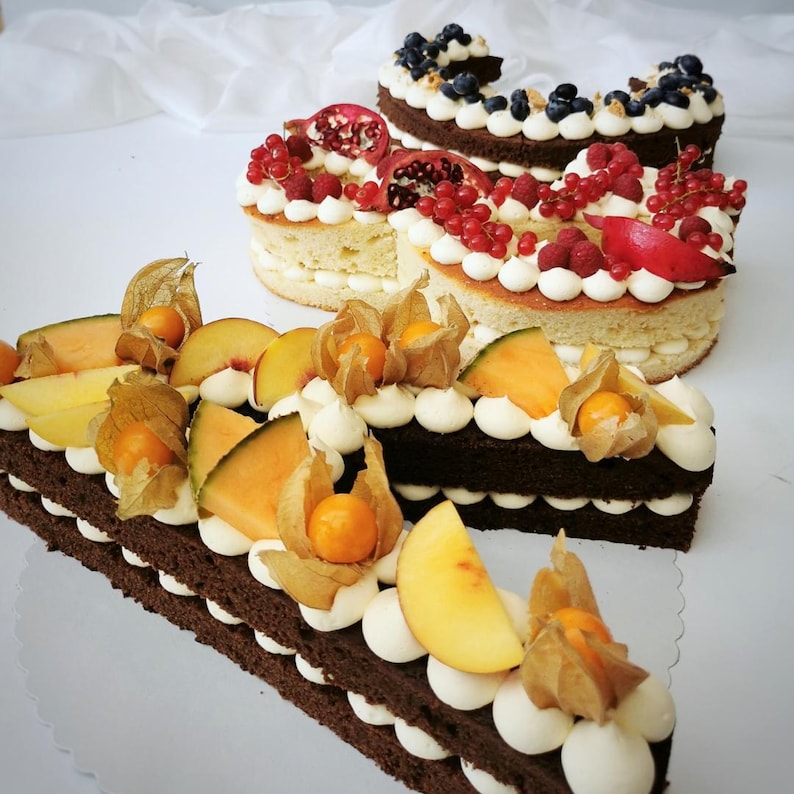 May include: Several letter-shaped cakes with white frosting and various fruit toppings. One cake is chocolate with melon, peach slices, and physalis. Other cakes have berries and pomegranate. The cakes are on a white surface.