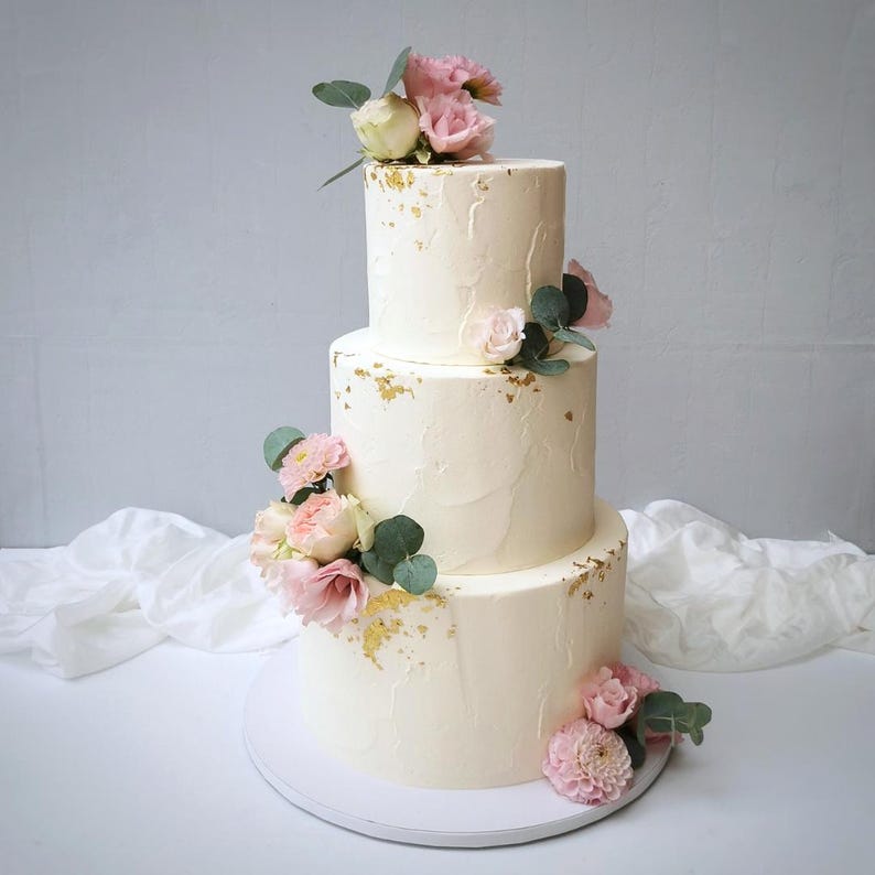 May include: A three-tiered white cake with textured frosting, adorned with pink and white flowers, green leaves, and gold leaf accents. The cake is on a white cake board, perfect for a wedding or celebration.