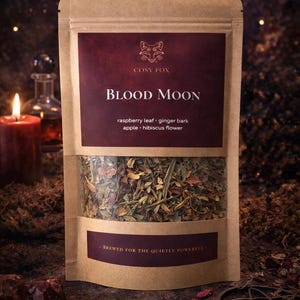 May include: A brown paper pouch of Cosy Fox Blood Moon tea with a clear window showing dried herbs. The label features a fox icon, the text Blood Moon, ingredients including raspberry leaf, ginger bark, apple, hibiscus flower, and the text Brewed for the quietly powerful.