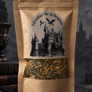 May include: A brown paper stand-up pouch with a clear window revealing a dried herbal tea blend. The label features a black and white illustration of a castle, two flying dragons, and the text "I'd rather be at Basgiath" in a curved font.