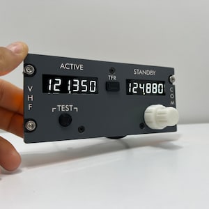 May include: A gray aircraft radio transceiver with a digital display showing "12.1350" and "12.4880". The transceiver has a "TEST" button, a "TFR" button, and a knob labeled "COM".