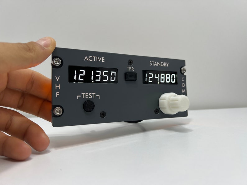 May include: A gray aircraft radio transceiver with a digital display showing "12.1350" and "12.4880". The transceiver has a "TEST" button, a "TFR" button, and a knob labeled "COM".