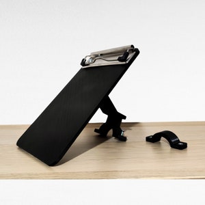 May include: A black clipboard with a metal clip is attached to a black stand. The stand has a metal bracket that can be used to attach the clipboard to a desk or table. A separate black metal bracket is also included.