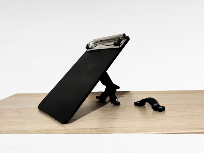 May include: A black clipboard with a metal clip is attached to a black stand. The stand has a metal bracket that can be used to attach the clipboard to a desk or table. A separate black metal bracket is also included.