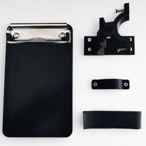 May include: A black clipboard with a silver clip and a black plastic mounting bracket, a black plastic handle, and a black plastic strap.
