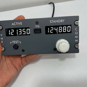 May include: A gray aircraft radio transceiver with a digital display showing "12.1350" and "124.880". The transceiver has a knob and a button labeled "TEST".