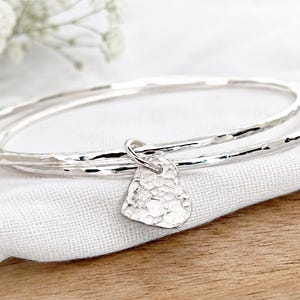 May include: Two silver bangles with a hammered texture, featuring a single heart-shaped charm attached to one of the bracelets. The metal surface of both the bangles and the heart charm reflects light, creating a bright, metallic appearance.