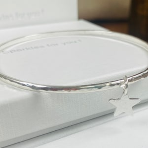 May include: A silver bangle bracelet with a hammered finish and a star charm. The bracelet is displayed in a white box with the words "Sparkles for you!" printed on the inside.