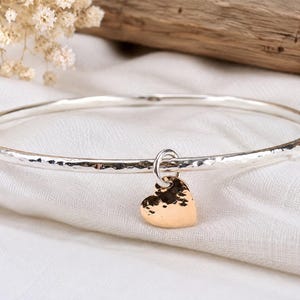 May include: A silver bangle bracelet with a hammered texture and a single small heart-shaped charm. The heart charm features a contrasting gold-toned finish and a matching hammered surface, suspended from the bangle by a small silver jump ring.