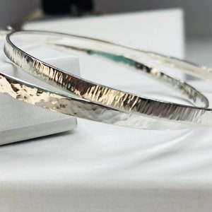 May include: Two silver hammered bangle bracelets, one slightly larger than the other, are stacked on top of each other on a white surface.