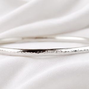 May include: A silver-toned bangle bracelet featuring a hammered, textured surface that creates a series of small, reflective indentations along the entire outer circumference of the circular metal band, showcasing a polished and metallic finish.