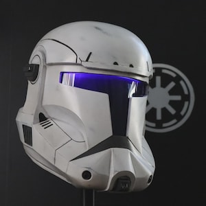 Pode incluir: Um capacete branco e preto com viseira azul, que lembra o capacete de um soldado espacial. O capacete tem uma aparência desgastada, com detalhes em preto e uma viseira proeminente. Um emblema branco se destaca em um fundo preto.