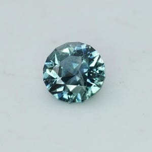 May include: A round, faceted gemstone with a teal and blue color. The gem has a brilliant cut, reflecting light and creating a sparkling effect. The gemstone is set against a white background, highlighting its color and clarity. This gem is suitable for jewelry.
