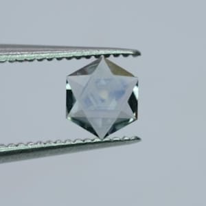 Puede incluir: Una gema clara y facetada sujeta con pinzas. La gema está tallada en forma de Estrella de David, con seis puntas y un cuerpo hexagonal. La gema tiene un ligero tono azul y parece transparente.