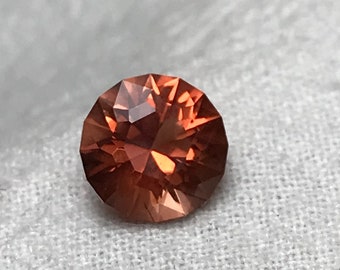 May include: A round, faceted, orange gemstone with a reddish hue.