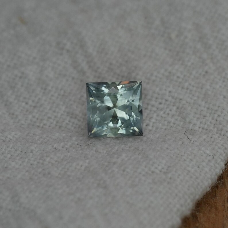 May include: A square-cut gemstone with a light sage green hue and a faceted surface that reflects light. The stone features a precise geometric shape with sharp corners and a brilliant, clear appearance, showcasing internal light refraction patterns.
