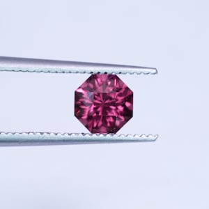 May include: A faceted, octagonal gemstone in a deep magenta hue is held by a pair of tweezers. The gem's cut reveals intricate internal facets, reflecting light. The tweezers are silver-colored, and the background is a soft white.