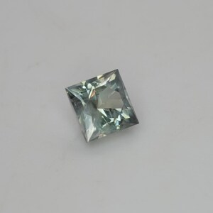 May include: A square-cut gemstone with a faceted surface, displaying a pale, translucent sage green color. The stone features a geometric princess cut with sharp corners and internal light reflections visible throughout the transparent crystalline structure.