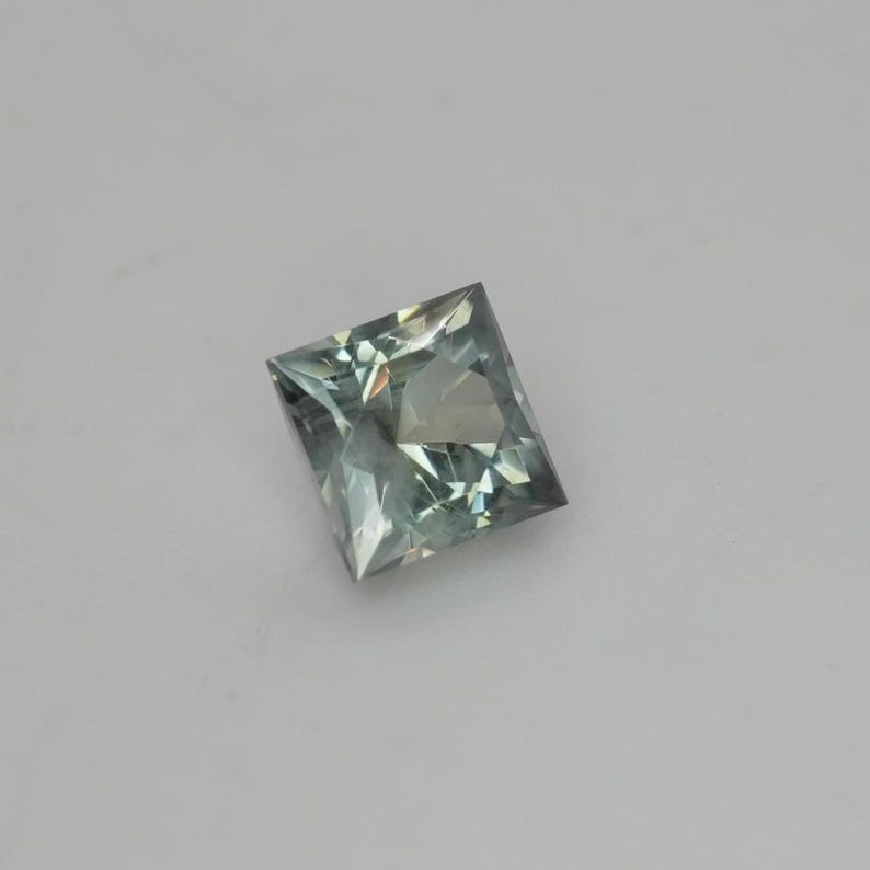 May include: A square-cut gemstone with a faceted surface, displaying a pale, translucent sage green color. The stone features a geometric princess cut with sharp corners and internal light reflections visible throughout the transparent crystalline structure.
