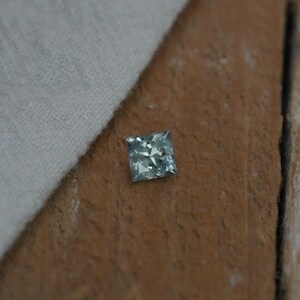 May include: A square-cut gemstone with a faceted surface, displaying a pale blue-green color. The stone features a geometric arrangement of triangular facets that reflect light, creating a sparkling appearance across its top surface.