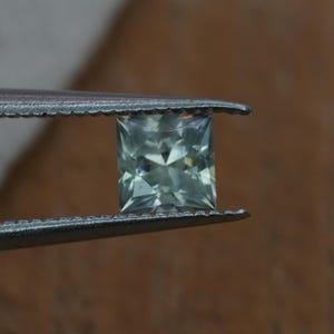 May include: A square-cut gemstone with a pale sage green hue and multifaceted surface held by metal tweezers. The stone features a clear, geometric arrangement of facets that reflect light, creating a sharp, symmetrical appearance.