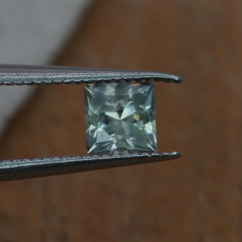 May include: A square-cut gemstone with a pale sage green hue and multifaceted surface held by metal tweezers. The stone features a clear, geometric arrangement of facets that reflect light, creating a sharp, symmetrical appearance.