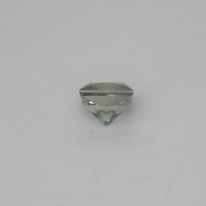 May include: A faceted, cushion-cut gemstone with a pale, translucent green hue. The stone features a symmetrical arrangement of triangular and trapezoidal facets that converge at a pointed base, creating a clear, reflective, and geometric appearance.