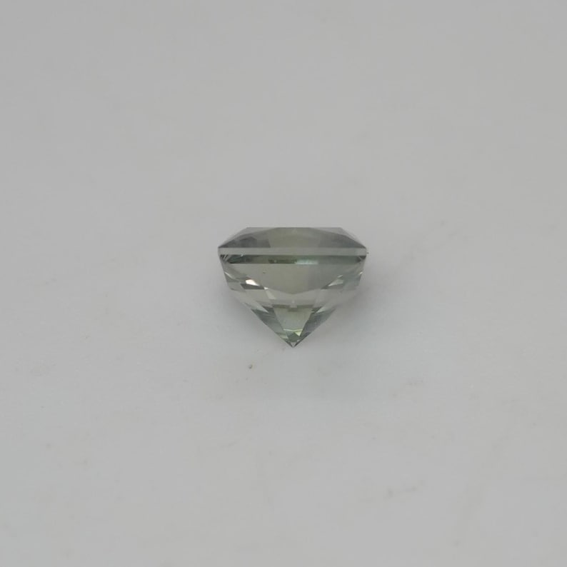 May include: A faceted, cushion-cut gemstone with a pale, translucent green hue. The stone features a symmetrical arrangement of triangular and trapezoidal facets that converge at a pointed base, creating a clear, reflective, and geometric appearance.