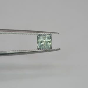 May include: A square-cut, pale mint green gemstone held by a pair of serrated metal tweezers. The stone features a faceted surface that reflects light, displaying a translucent quality with subtle variations in its light green color throughout.