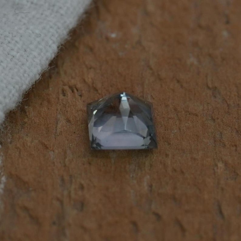 May include: A square-cut gemstone with a faceted surface and a translucent gray color. The stone features a geometric shape with multiple triangular facets that reflect light, creating a sharp, angular appearance across its top surface.