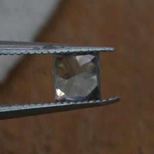 May include: A square-cut, translucent gemstone with a faceted surface held by a pair of metal tweezers. The stone displays a pale, neutral color with light reflecting off its geometric facets, showcasing a clear and polished appearance.