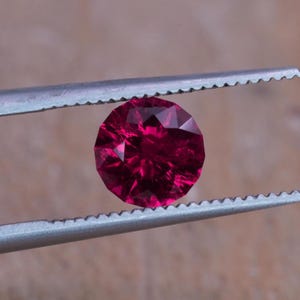Op de afbeelding: Een gefacetteerde, ronde, dieprode edelsteen wordt vastgehouden door een zilveren pincet. De edelsteen heeft meerdere facetten die het licht reflecteren, waardoor een sprankelend effect ontstaat. De achtergrond is een vage, neutrale tint.