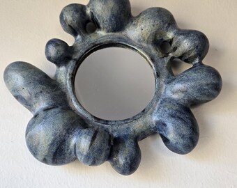 May include: A unique, handcrafted wall mirror with a round reflective surface. The frame is an organic shape with a blue and gray marbled finish. The mirror is a decorative piece for home decor.
