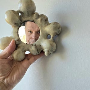 May include: A distinctive, beige-toned mirror with an abstract, flower-like design. The mirror is held in a hand, reflecting a person's face. The frame has a textured, irregular surface, enhancing its artistic quality.